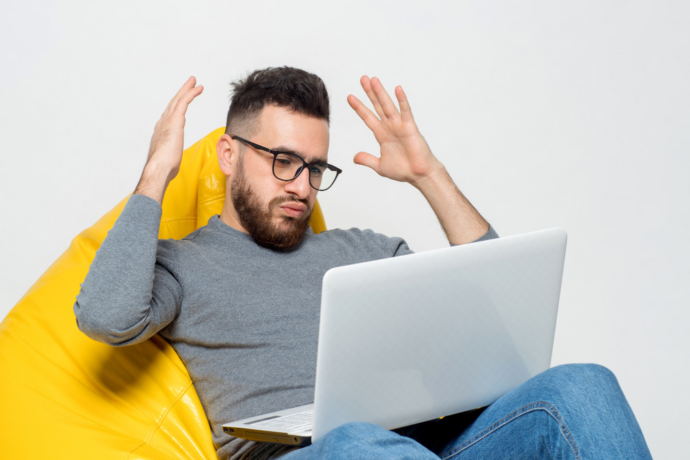 Photo for 5 Website Mistakes That Are Costing You Sales blog post. A young man sits on a bright yellow beanbag, with a laptop on his lap and he holds up both of his hand, showing that he had enough and he is fed up.